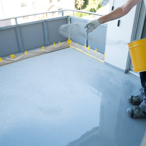 Balkonsanierung: Handwerker trägt frische Epoxid-Beschichtung auf Balkonboden auf und streut Sand für rutschfeste Oberfläche, schützt vor Feuchtigkeit und Frostschäden